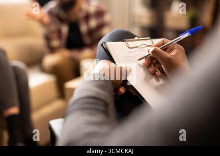 Primo piano delle mani afroamericane che scrivono con penna su carta, documentando la sessione in ufficio. Terapeuta maschile di colore seduto con appunti, prendendo appunti attentamente durante la consultazione. Foto Stock
