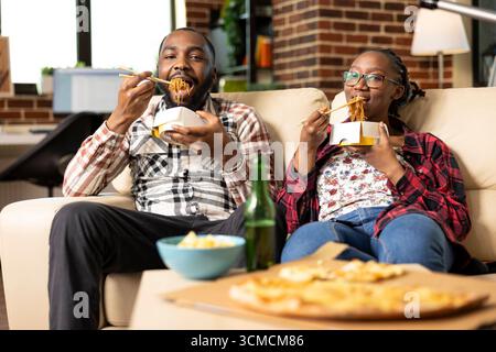 Giovani coppie che si rilassano sul divano del soggiorno, usano le bacchette per mangiare spaghetti dal takeout box e guardano la televisione. Uomo e donna di colore si legano all'intrattenimento e al cibo in casa. Foto Stock