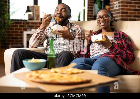 Una giovane coppia nera si rilassa a casa, guardando la televisione insieme al cibo da asporto. Fidanzato afroamericano che mangia spaghetti con la fidanzata, si gode il tempo libero in un appartamento con pareti in mattoni. Foto Stock