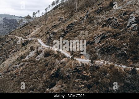 Strada sterrata stretta e tortuosa che attraversa ripidi pendii rocciosi ricoperti da arbusti secchi, alberi sparsi e formazioni rocciose aspre sotto il nuvoloso sk Foto Stock