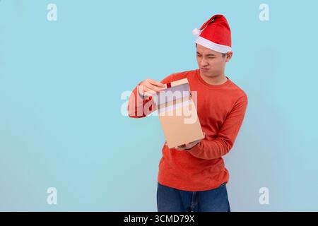 Un giovane dall'espressione delusa che indossa un cappello di Babbo Natale e un maglione rosso scopre una piccola scatola regalo marrone su sfondo blu Foto Stock