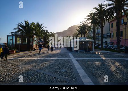 Camminando lungo la strada lastricata principale, la passeggiata sulla spiaggia, alla luce del sole nel tardo pomeriggio. Nella città balneare di Finalmarina, in Liguria. Foto Stock