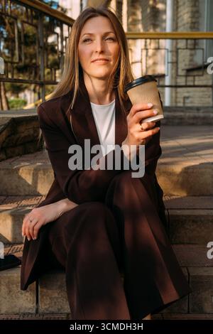 Ritratto urbano. Caffè diurno. Donna d'affari fiduciosa e penosa che si diverte da sola con una bevanda calda in tazza di carta. Foto Stock
