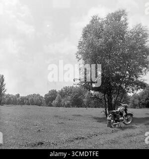 Questa suggestiva fotografia d'archivio in bianco e nero degli anni '1970 trasmette un senso di libertà e solitudine nella campagna sovietica. Un'iconica motocicletta sovietica IZH è parcheggiata sotto un albero solitario in un grande campo sullo sfondo di una foresta. Catturata nella regione di Donec'k della RSS Ucraina, la scena riflette il romanticismo dei viaggi semplici e la popolarità delle motociclette come simbolo della mobilità personale durante l'"era della stagnazione". È un'immagine nostalgica di una fuga estiva, un legame con la natura e la cultura del tempo libero in URSS Foto Stock