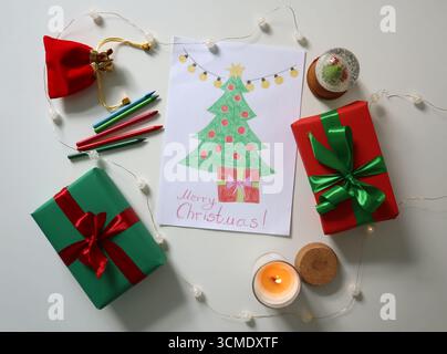 Disegno per bambini di un albero di Natale decorato con un regalo sotto. Circondato da regali di Natale avvolti di colore verde, rosso con archi, luci a corda, candele accese, matite colorate. Sfondo bianco. Foto Stock