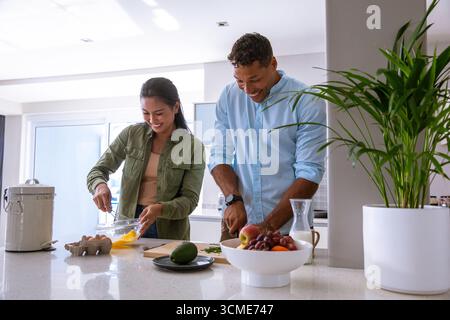 Coppia variegata che si trova sull'isola bianca della cucina, sbattere le uova in un recipiente di vetro, tritare le erbe Foto Stock