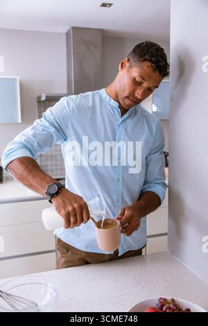Uomo che versa il latte dal biberon di vetro alla tazza in ceramica rosa sul piano di lavoro della cucina accanto al recipiente per mescolare Foto Stock