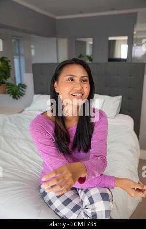 Donna asiatica di mezza età seduta sul letto, che si allontana sorridendo in camera da letto con piante in vaso Foto Stock