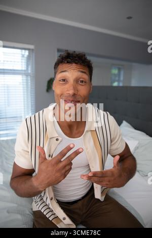 Uomo seduto sul letto in camera da letto, indossa camicia a righe accanto alla testiera e alle tende alle finestre Foto Stock