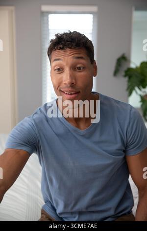 Uomo seduto sul letto accanto alle tende alle finestre e piante in vaso che parlano fuori dalla telecamera nella camera da letto grigia Foto Stock