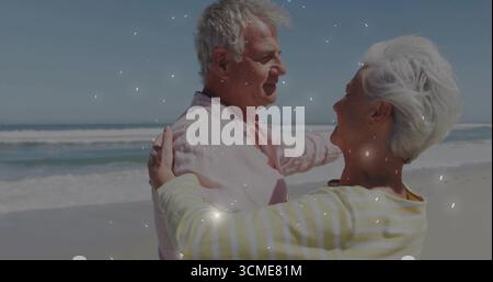 Abbracciare una coppia senior in piedi sulla spiaggia di sabbia, sorridere con camicia rosa pastello e top a righe Foto Stock