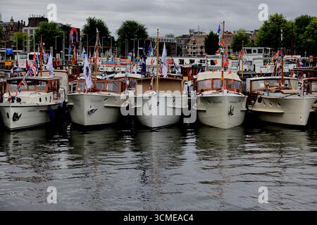 Fila di berline d'epoca decorate con bunting per la Vela 2025, ormeggiate fianco a fianco ad Amsterdam, Paesi Bassi, riflessi sull'acqua sotto il coperto. Foto Stock