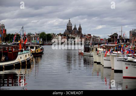 Linea di barche classiche decorate di fronte al centro storico durante la navigazione 2025 sull'IJ di Amsterdam, Paesi Bassi, riflessi sotto il cielo coperto. Foto Stock
