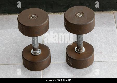 Primo piano di un set di manubri in metallo ben usato con ruggine e graffi visibili, appoggiato sul pavimento della palestra, simboleggia il duro lavoro e il fitness vintage. Foto Stock