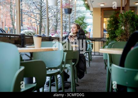 Donna di mezza età con cappotto invernale che gusta un caffè con panna montata presso un caffè all'aperto innevato, atmosfera natalizia, stile di vita urbano accogliente. Foto Stock