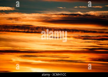 Una vista ravvicinata e astratta di un cielo al tramonto pieno di nuvole vivaci e dinamiche. I colori variano da arance profonde e oro a rossi e marroni tenui, cr Foto Stock