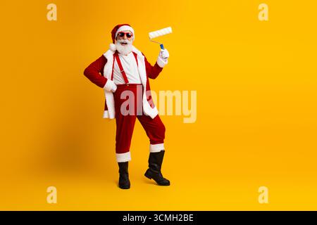 Babbo Natale indossato con abiti festivi tenendo in mano un rullo di pittura, posato energicamente su uno sfondo giallo brillante Foto Stock