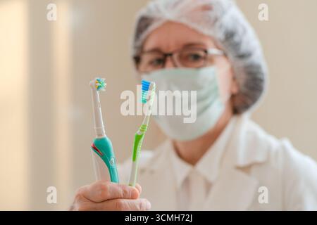 Primo piano di una dentista in uniforme bianca con uno spazzolino manuale e uno elettrico. Concentrati sulle sue mani. Promuovere una buona igiene orale e cure odontoiatriche. Scelta tra gli strumenti di pulizia. Foto Stock