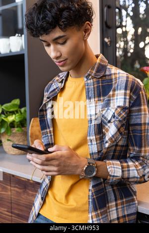 Uomo in camicia a quadri in piedi in cucina a controllare lo smartphone accanto a tazze e piante in ceramica Foto Stock