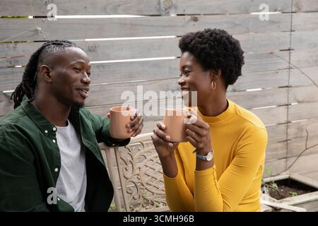 Coppia afroamericana che indossa la parte superiore della senape seduta sulla panchina nel cortile che regge tazze Foto Stock