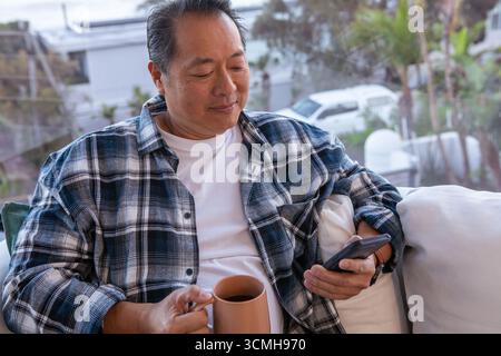 Uomo cinese anziano seduto su un divano imbottito vicino alla finestra, tenendo in mano smartphone e tazza da caffè Foto Stock