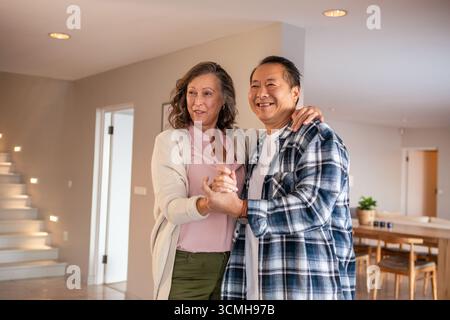 Diverse coppie senior che ballano lentamente in un'area soggiorno open space con scala illuminata e tavolo da pranzo Foto Stock