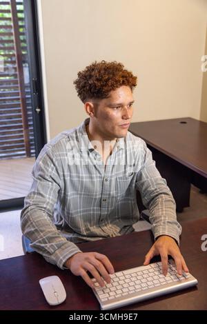 Maschio adulto che digita la tastiera wireless su una scrivania in legno facendo scorrere la porta in vetro in casa Foto Stock