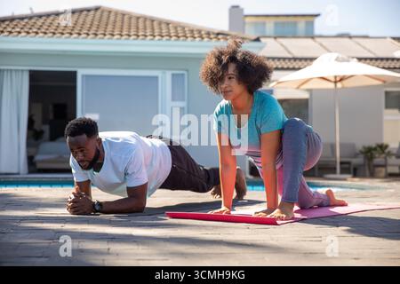 Diverse coppie che si allenano su stuoie yoga accanto alla piscina sul patio in pietra sotto l'ombrellone del patio Foto Stock