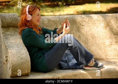 Donna felice con le cuffie che ascolta la musica e usa lo smartphone mentre è seduta sulla panchina in un parco Foto Stock