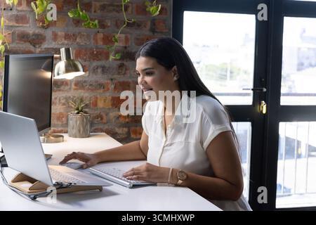 Donna adulta di medie dimensioni che digita con tastiera wireless su una scrivania bianca con laptop, spazio di copia Foto Stock