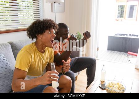 Diversi amici maschi seduti su un divano grigio nel soggiorno mangiano popcorn e bevono bibite tifose Foto Stock