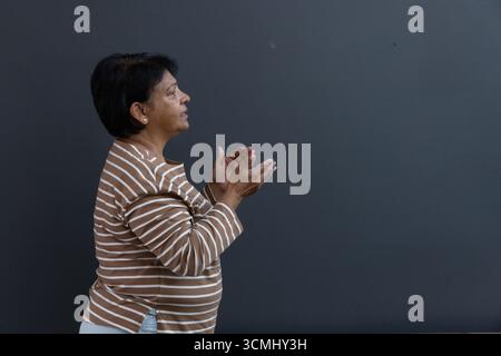 Donna indiana anziana che indossa una camicia a righe che stringe le mani davanti alla parete grigia, copia spazio Foto Stock