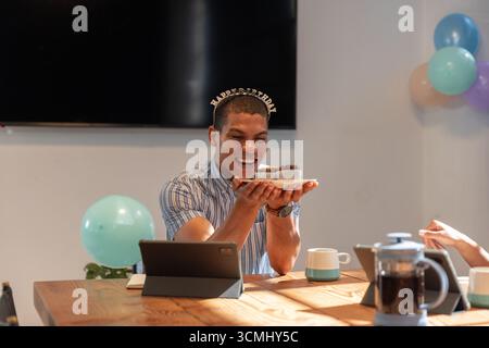 Uomo afro-americano seduto al tavolo delle riunioni tenendo una torta di compleanno, una candela accesa in mezzo a palloncini Foto Stock