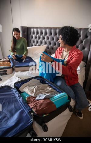Coppia afroamericana e indiana che imballano vestiti, maschera facciale bianca in valigie aperte sul letto Foto Stock