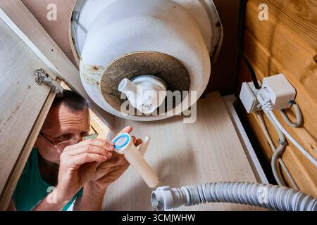 Handyman installa una rondella in gomma tra il tubo di uscita e il tubo di collegamento del pozzetto di raccolta bottiglie lavorando in spazi ristretti sotto il lavandino. Foto Stock