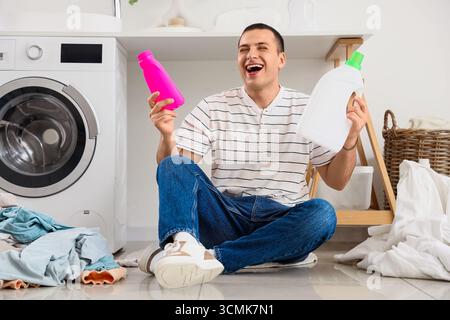 Giovane felice con bottiglie di detergente e pile di vestiti sporchi seduti sul pavimento nella lavanderia Foto Stock