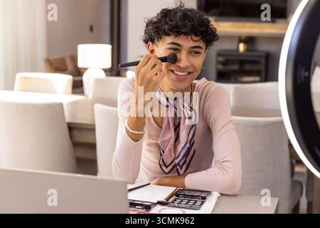 Uomo seduto nella sala da pranzo che applica il trucco con tavolozze di pennelli e illuminatore anulare Foto Stock