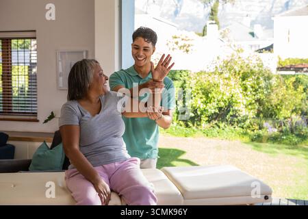Terapeuta maschile che guida la donna anziana attraverso il braccio allungato su un lettino di trattamento imbottito in camera Foto Stock