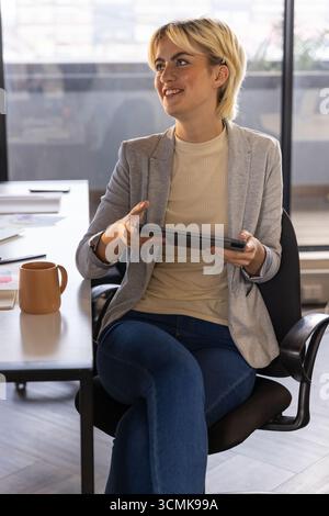 Donna professionista con un blazer che tiene un tablet, rivede i documenti alla scrivania con la tazza Foto Stock