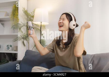 Una giovane donna ama giocare ai videogiochi a casa, indossare le cuffie e tenere in mano un controller con entusiasmo. Si siede comodamente su un divano, sorridendo Foto Stock