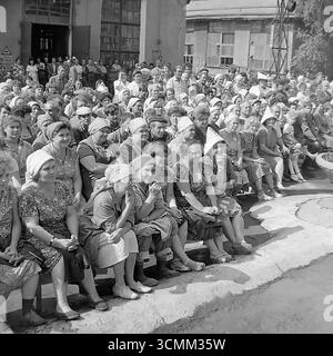Questa fotografia d'archivio espressiva degli anni '1970-'1980 cattura il pubblico dei lavoratori in un concerto improvvisato nel cortile dell'armatura e dell'isolatore di Slavyansk (AIZ) nella RSS Ucraina. Un grande gruppo di persone, per lo più donne nei loro abiti da lavoro quotidiani, guarda lo spettacolo con genuino interesse e attenzione. L'immagine è un potente documento sociale, che ritrae realisticamente la classe operaia sovietica e la pratica culturale degli "incontri creativi", dove l'arte veniva portata direttamente alle imprese industriali Foto Stock