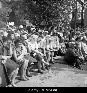 Questa fotografia d'archivio espressiva degli anni '1970-'1980 cattura il pubblico dei lavoratori in un concerto improvvisato nel cortile dell'armatura e dell'isolatore di Slavyansk (AIZ) nella RSS Ucraina. Un grande gruppo di persone, per lo più donne nei loro abiti da lavoro quotidiani, guarda lo spettacolo con genuino interesse e attenzione. L'immagine è un potente documento sociale, che ritrae realisticamente la classe operaia sovietica e la pratica culturale degli "incontri creativi", dove l'arte veniva portata direttamente alle imprese industriali Foto Stock