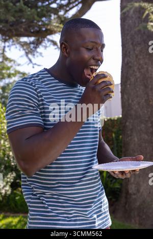 Uomo afro-americano che morde l'hamburger tenendo un piatto di carta nel cortile posteriore con indosso una camicia Foto Stock Uomo afro-americano che morde l'hamburger tenendo un piatto di carta nel cortile posteriore con indosso una camicia Foto Stock