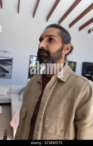 Uomo indiano medio adulto che indossa una camicia beige e una t-shirt ruggine in piedi nel soggiorno accanto al divano Foto Stock