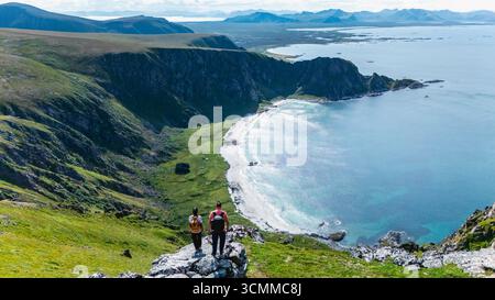 Due escursionisti godono di viste mozzafiato dal monte Matind mentre guardano verso il basso la pittoresca spiaggia di Bleik e il vibrante paesaggio artico di Andoya, Norvegia, in una limpida giornata estiva. Foto Stock