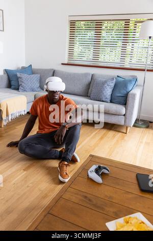 Uomo afro-americano che indossa un visore vr che regge il controller vr sul pavimento in salotto Foto Stock