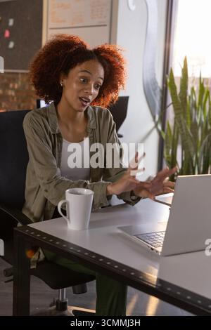 Donna afroamericana che gestiva mentre parla alla scrivania in ufficio con un computer portatile e una tazza da caffè Foto Stock