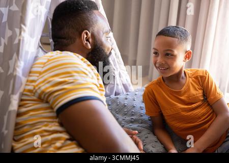 Padre e figlio afroamericani che giocano all'interno della tenda in tessuto stellare a casa su cuscino grigio Foto Stock