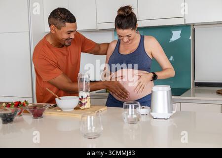 Diverse coppie che si aspettano di mettere le mani sulla pancia incinta nella cucina di casa con ciotole e frullatore Foto Stock