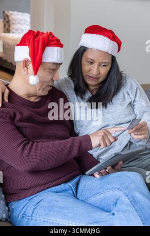 Coppia senior in cappelli di babbo natale che naviga online su tablet con carta di credito in salotto Foto Stock